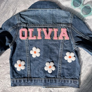 May include: A blue denim jacket with white daisy patches and a pink felt name patch that says "OLIVIA".