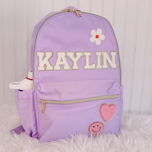 May include: A lavender backpack with gold zippers and the name "KAYLIN" in white letters with gold trim. The backpack has a white flower patch, a pink heart patch, and a smiley face patch. A water bottle is in the side pocket.