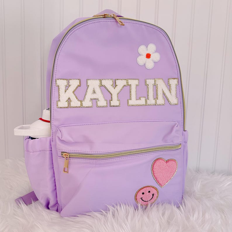Pu&ograve; includere: Uno zaino lavanda con cerniere dorate e il nome "KAYLIN" in lettere bianche e dorate. Presenta una toppa a forma di fiore bianco, un cuore rosa e una toppa con faccina sorridente. Una bottiglia d'acqua &egrave; nella tasca laterale.