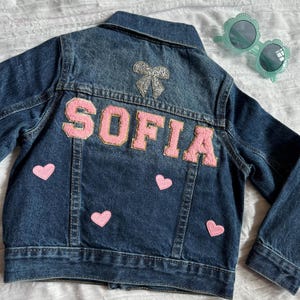 May include: A dark blue denim jacket with the name "SOFIA" in pink letters on the back. The jacket has a silver bow and pink heart patches. A pair of light green sunglasses with a floral design are also in the image.