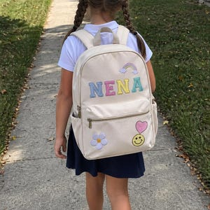 Pu&ograve; includere: Uno zaino beige con il nome "NENA" ricamato in colori pastello, un arcobaleno, un fiore, un cuore e una faccina sorridente. Lo zaino ha una tasca frontale con cerniera e tasche laterali. La bambina indossa una camicia bianca, una gonna blu navy e scarpe da ginnastica rosa.