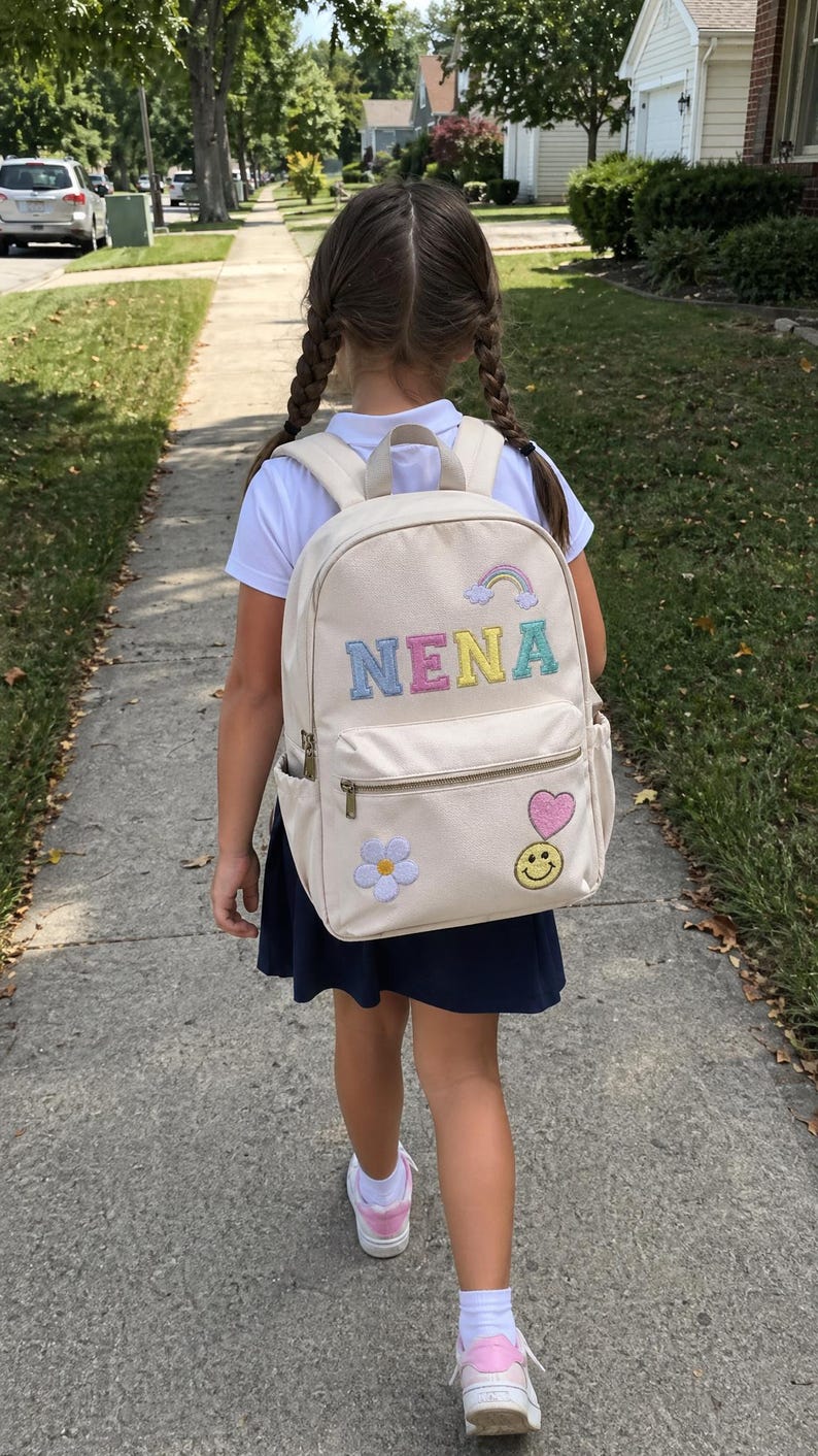 Pu&ograve; includere: Uno zaino beige con il nome "NENA" ricamato in colori pastello, un arcobaleno, un fiore, un cuore e una faccina sorridente. Lo zaino ha una tasca frontale con cerniera e tasche laterali. La bambina indossa una camicia bianca, una gonna blu navy e scarpe da ginnastica rosa.