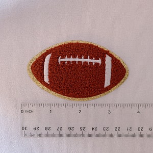 Op de afbeelding: Een geborduurde voetbalpatch in bruin en wit met een gouden rand. De patch is ongeveer 5 cm in diameter.