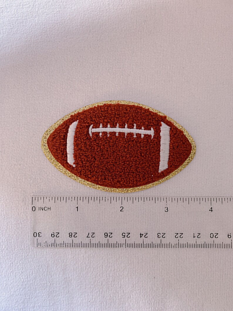 Op de afbeelding: Een geborduurde voetbalpatch in bruin en wit met een gouden rand. De patch is ongeveer 5 cm in diameter.