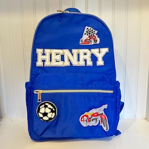 Pu&ograve; includere: Uno zaino blu reale con cerniere dorate e il nome "HENRY" in lettere bianche e dorate. Le toppe includono un'auto da corsa, un pallone da calcio e una motocicletta rossa. Adatto per trasportare materiale scolastico o oggetti personali.