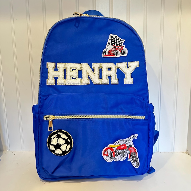 Pu&ograve; includere: Uno zaino blu reale con cerniere dorate e il nome "HENRY" in lettere bianche e dorate. Le toppe includono un'auto da corsa, un pallone da calcio e una motocicletta rossa. Adatto per trasportare materiale scolastico o oggetti personali.