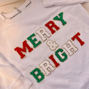 May include: White sweatshirt with the words "Merry & Bright" in red, green, and gold letters. The letters are outlined in gold and have a fuzzy texture. The sweatshirt has a Calvin Klein Performance logo on the collar.