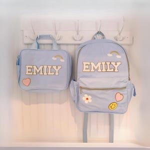 Peut inclure: Un sac à déjeuner bleu clair et un sac à dos assorti, tous deux portant le nom EMILY en lettres blanches de style universitaire avec une bordure dorée. Les sacs sont ornés d'écussons brodés d'un arc-en-ciel, d'un cœur rose, d'une fleur blanche et d'un smiley jaune.