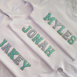 May include: Three white sweatshirts with different names embroidered on them in gold and light blue lettering. The names are "Jakey", "Jonah", and "Myles".