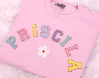 May include: Pink sweatshirt with the name "PRISCILLA" spelled out in colorful, glittery letters in an arc. A white flower with a red center is below the name.
