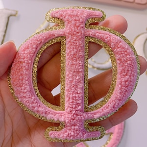 May include: A pink, textured Greek letter Phi patch with gold glitter trim. The patch is held in a hand, with a blurred background of other patches and a pink keyboard.