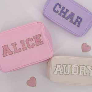 May include: Three rectangular cosmetic bags in pastel colors: pink, lavender, and cream. Each bag features a name in a varsity-style font with gold glitter accents. Pink heart-shaped patches are scattered around the bags.