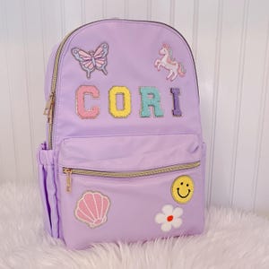 May include: A lavender backpack with gold zippers and patches. Patches include a butterfly, unicorn, seashell, flower, and a smiley face. The name "CORI" is spelled out in colorful, sequined letters on the front. The backpack has a front pocket and side pocket.