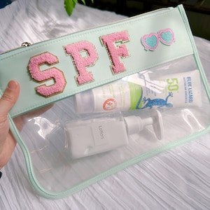 May include: A light blue clear plastic zippered pouch with the word "SPF" in pink and gold letters. The pouch contains two bottles of sunscreen, one with the text "Blue Lizard Australian Sunscreen 50+" and the other with the text "Lotion".