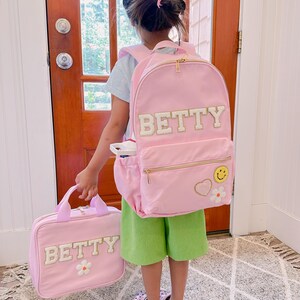 Pu&ograve; includere: Uno zaino rosa e una borsa per il pranzo abbinata con il nome "BETTY" a lettere bianche. Lo zaino ha una cerniera dorata e una toppa a forma di faccina sorridente. La borsa per il pranzo ha una toppa a forma di fiore bianco. Il bambino indossa pantaloncini verdi.
