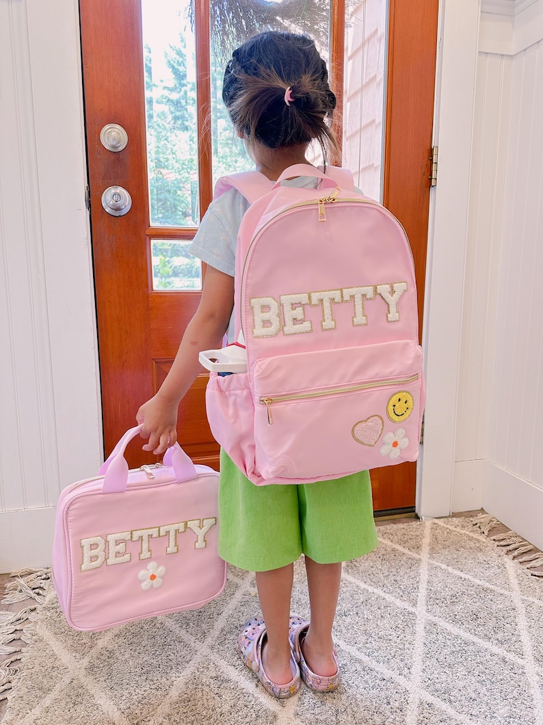 Pu&ograve; includere: Uno zaino rosa e una borsa per il pranzo abbinata con il nome "BETTY" a lettere bianche. Lo zaino ha una cerniera dorata e una toppa a forma di faccina sorridente. La borsa per il pranzo ha una toppa a forma di fiore bianco. Il bambino indossa pantaloncini verdi.