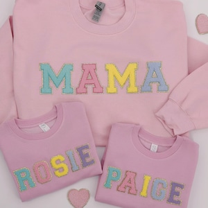 May include: Three pink sweatshirts with colorful glitter lettering. The largest sweatshirt says "MAMA" and the two smaller sweatshirts say "ROSIE" and "PAIGE".