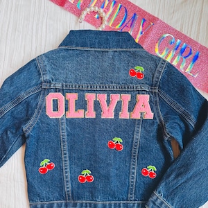 May include: A blue denim jacket with pink felt lettering that says "OLIVIA" and several red cherry patches.