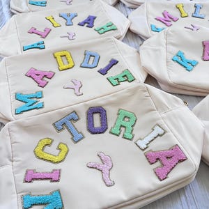 May include: A collection of off-white canvas bags with colorful embroidered letters spelling out names. The bags are arranged in a pile, with the top bag showing the name "VICTORIA" in a rainbow of colors.