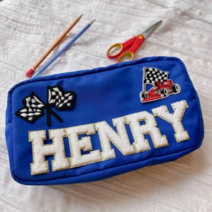 Peut inclure: Une trousse bleue avec le nom "HENRY" en lettres blanches et duveteuses avec une bordure dorée. La trousse présente des écussons sur le thème de la course, notamment des drapeaux à damiers et une voiture de course rouge. Un crayon et un stylo sont à côté de la trousse, ainsi que des ciseaux rouges.