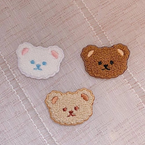 May include: Three embroidered felt bear patches in white, brown, and beige. The bears have black eyes and noses and are smiling.