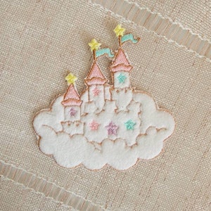 May include: A white felt patch with an embroidered pink castle on a cloud. The castle has multiple towers and flags with stars. The cloud has pink, purple, and green stars.