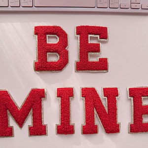May include: Red felt letters spelling out "BE MINE" with gold glitter trim. The letters are arranged in a line on a pink background.