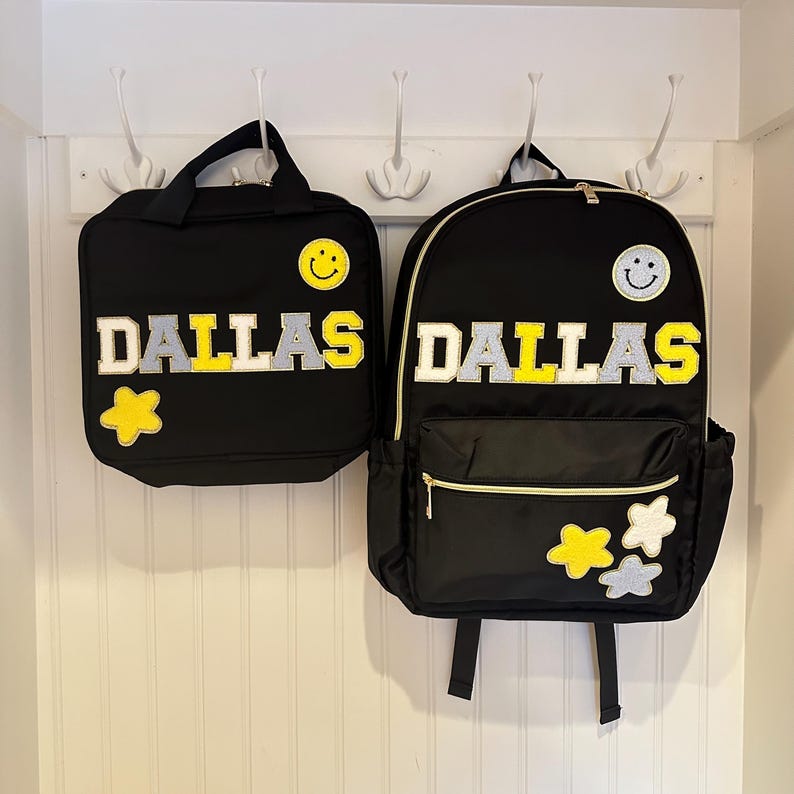 Pu&ograve; includere: Set di zaino e borsa per il pranzo neri con il nome "DALLAS" in lettere bianche, grigie e gialle. Entrambi gli articoli presentano una faccina sorridente e decorazioni floreali. Lo zaino ha una tasca frontale e tasche laterali.