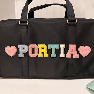 May include: Black duffel bag with a personalised name "PORTIA" in colourful felt letters and pink heart accents.