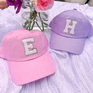 May include: Two baseball caps, one pink and one lavender, are displayed. Each cap features a large, white letter with a gold glitter outline. The pink cap has the letter "E", and the lavender cap has the letter "H".