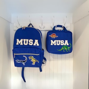 May include: Blue backpack and lunch bag set with the name "MUSA" in white block letters. The backpack has a front pocket and dinosaur patches. The lunch bag has a handle and dinosaur patches. Both bags are hanging on a white wall.