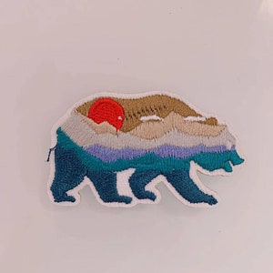 May include: Embroidered iron-on patch featuring a blue bear silhouette with a mountain range and a red sun in the background.