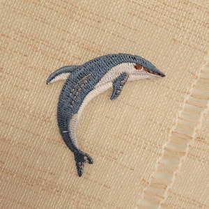 May include: Embroidered patch featuring a detailed dolphin design. The dolphin is primarily blue with white accents, and a touch of brown on the snout. The patch is set against a textured beige background, suggesting a nautical or marine theme.
