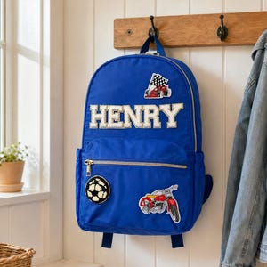May include: A blue backpack with the name "HENRY" in white block letters. Patches include a race car, a soccer ball, and a motorcycle. The backpack hangs on a wooden rack with a denim jacket to the side.
