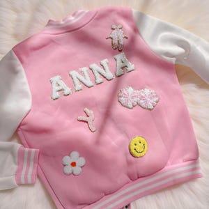 May include: Pink satin bomber jacket with white sleeves and trim. The jacket features the name "ANNA" in white, embellished letters, and various embroidered patches, including ballet slippers, pom-poms, a flower, and a smiley face.