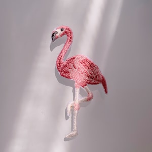 May include: Pink flamingo embroidered patch with white stitching. The flamingo is standing on one leg with its head turned to the side.