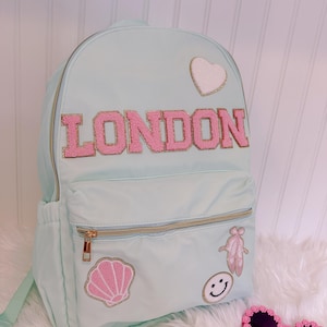 Pu&ograve; includere: Zaino azzurro con la scritta "LONDON" in lettere rosa. Lo zaino presenta toppe a forma di cuore, conchiglia, scarpette da ballo e faccina sorridente. Ha cerniere dorate.