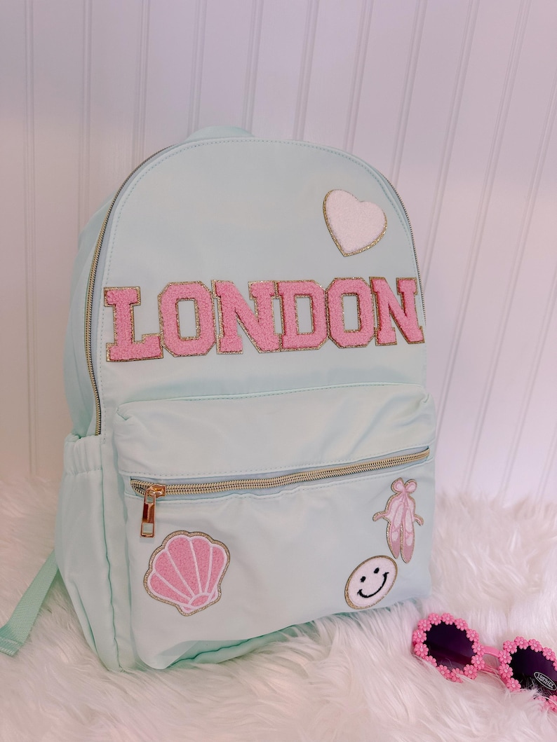 Pu&ograve; includere: Zaino azzurro con la scritta "LONDON" in lettere rosa. Lo zaino presenta toppe a forma di cuore, conchiglia, scarpette da ballo e faccina sorridente. Ha cerniere dorate.