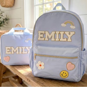May include: A light blue backpack and matching lunch bag set. Both items feature the name "EMILY" in gold-colored, block-letter patches. The backpack has a rainbow, flower, heart, and smiley face patch. The lunch bag has a rainbow and heart patch.