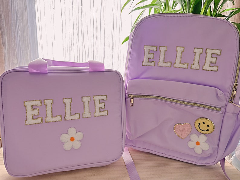 Pu&ograve; includere: Un set coordinato di zaino e borsa per il pranzo color lavanda. Entrambi gli articoli presentano il nome "ELLIE" in lettere bianche e dorate. Lo zaino ha una tasca frontale, cerniere dorate e toppe decorative, tra cui un cuore, una faccina sorridente e un fiore.