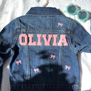 May include: A blue denim jacket with the name "OLIVIA" in pink letters on the back. There are five pink bows sewn on the jacket.