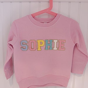 May include: Pink sweatshirt with the name "SOPHIE" spelled out in colorful, glittery letters.