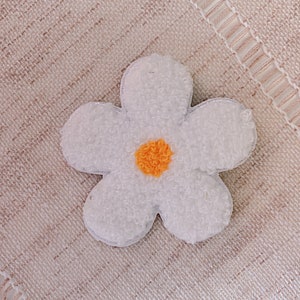 May include: A white fuzzy flower patch with a yellow center. The flower is on a beige textured background.