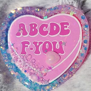 May include: A heart-shaped resin coaster with the text "ABCDE F-YOU" in white. The coaster is pink with a white border and filled with glitter, stars, and small beads. A small heart-shaped charm is also included.
