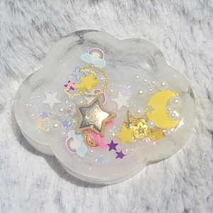 May include: Cloud-shaped resin art piece with a dreamy theme. The clear resin encases miniature stars, rainbows, and a yellow crescent moon. The piece is adorned with gold accents and iridescent glitter, creating a whimsical aesthetic.
