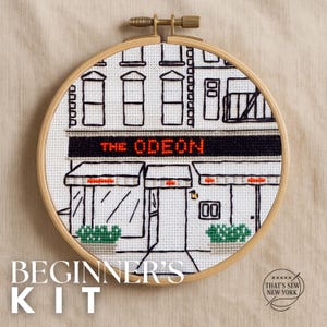 May include: A cross-stitch embroidery hoop featuring a black and white illustration of a building with the text "THE ODEON" in red. The words "BEGINNER'S KIT" are at the bottom. The embroidery is set in a wooden hoop.