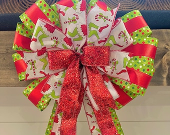 May include: A large Christmas bow featuring a Grinch-themed design. The bow has layers of red, green, and white ribbons. The white ribbon has a pattern of the Grinch's legs and candy canes. A sparkly red bow is in the center.
