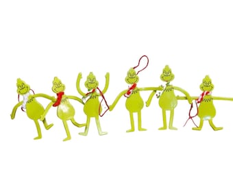 Op de afbeelding: Een set van zes limoengroene, platte, karaktervormige ornamenten die in een rij met elkaar verbonden zijn. Elk figuur heeft een puntig hoofd, een ondeugende gezichtsuitdrukking en dunne ledematen. Enkele figuren dragen kleine, kleurrijke wollen sjaals.