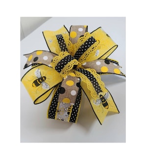 May include: A decorative bow featuring yellow and black ribbons with a bee theme. The bow has a mix of polka dots, bee illustrations, and lace trim. The color palette includes yellow, black, and brown.