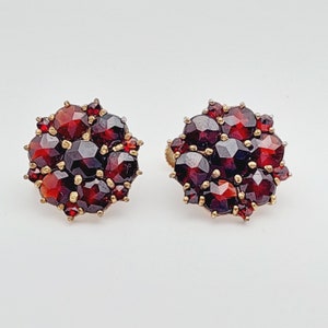 May include: A pair of gold earrings with a cluster of red garnet stones. The earrings are round and have a floral design.